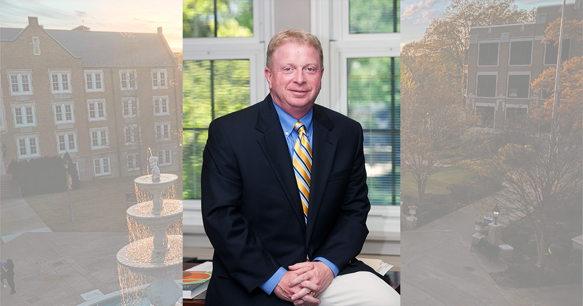 Portrait photo of College of Business and Technology Dean Greg Carnes sitting on the edge of his desk with his hands folded.