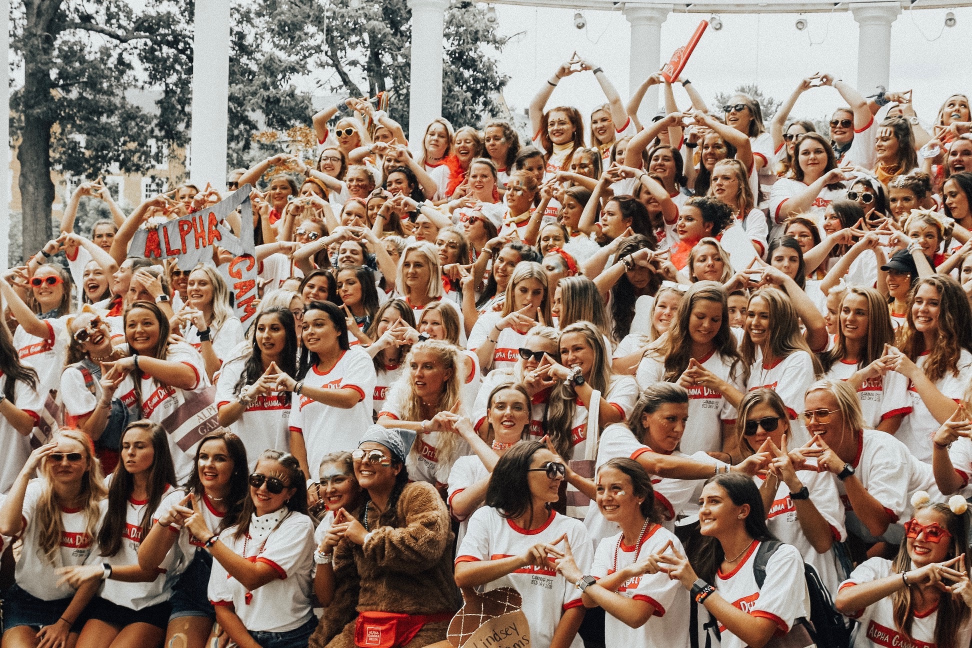 Women in white Alpha Gamma Delta shirts on bid day at the amphitheater posed holding their hands to form the sign of their sorority.